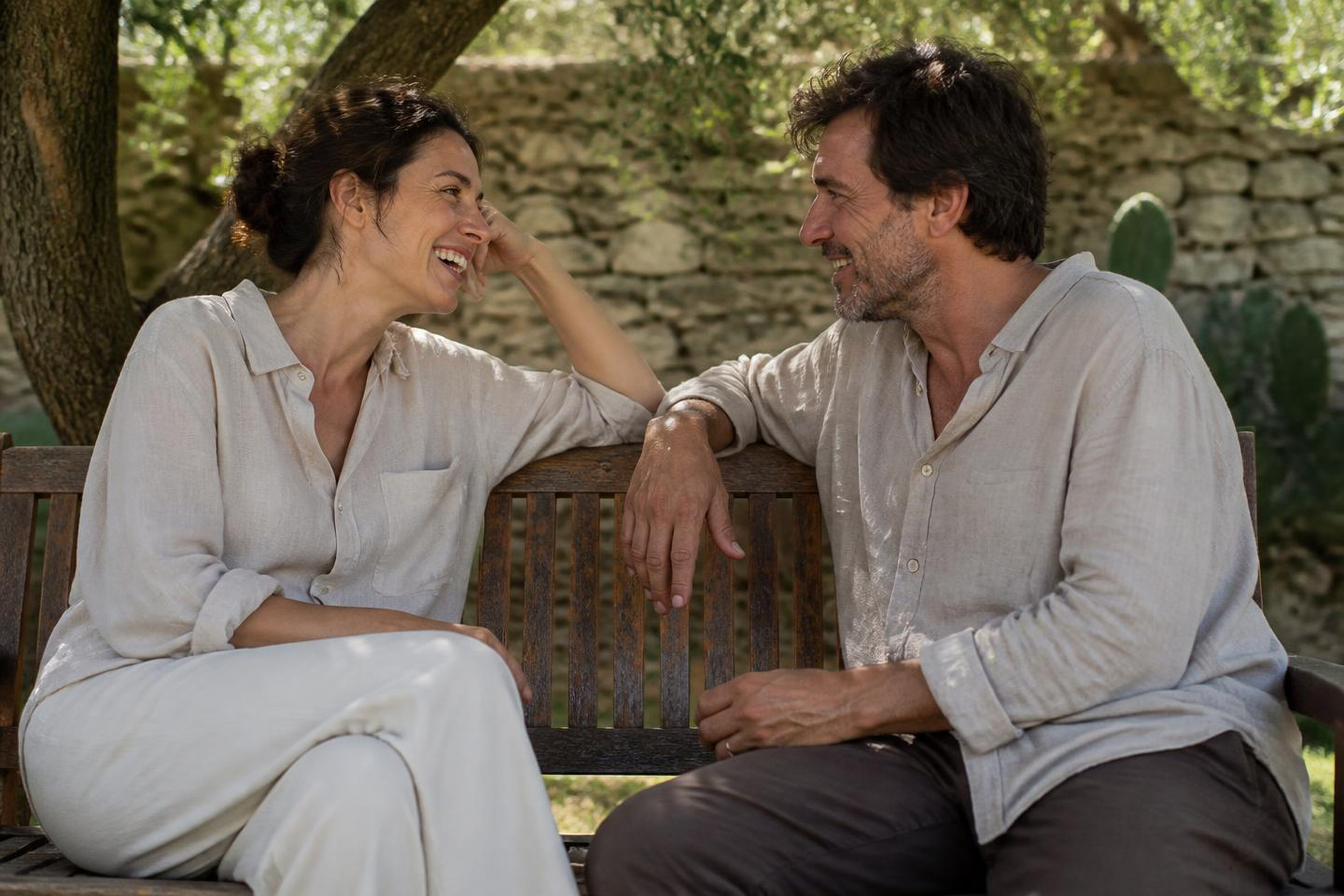 Two people laughing together on a garden bench under an olive tree